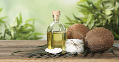 Coconut oil bottle, coconuts, and a jar of salt on a wooden surface with a green leafy background.