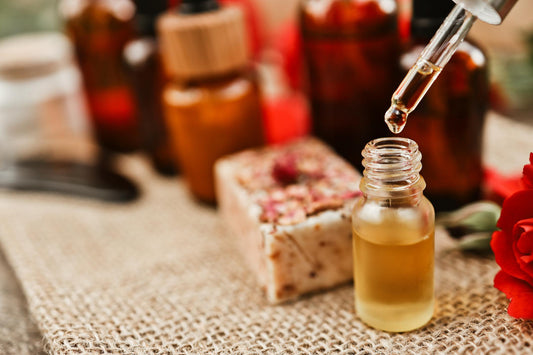 Dropper dispensing golden essential oil into a small glass bottle with rose petals and amber bottles in background